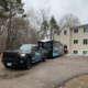 A moving truck and trailer parked outside of an apartment building.