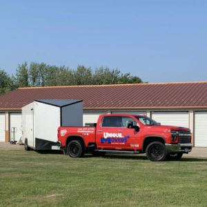 A red Unique Movers moving truck with a white moving trailer parked outside a storage unit.